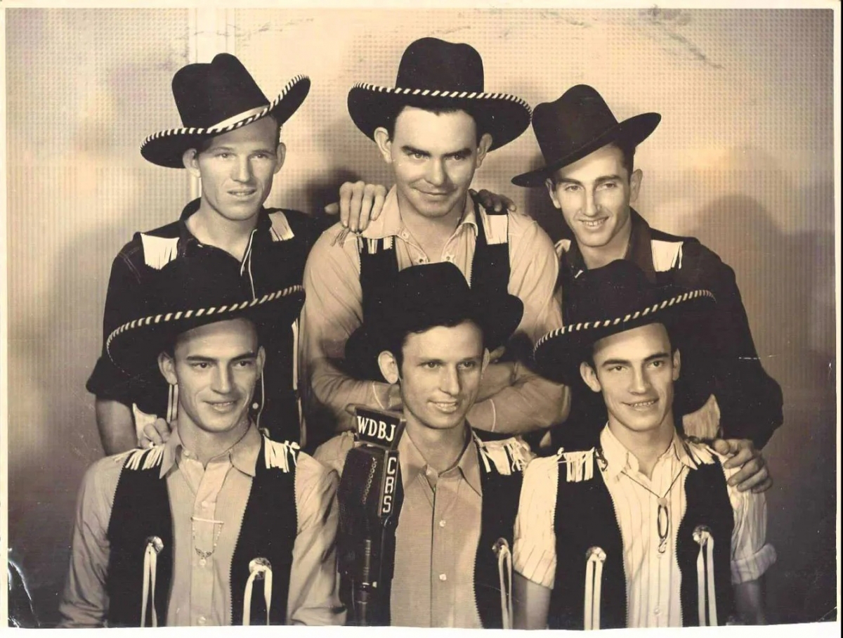 Sepia image of six smiling men standing in two rows of three in front of a microphone. Wearing Western wear and cowboy hats.