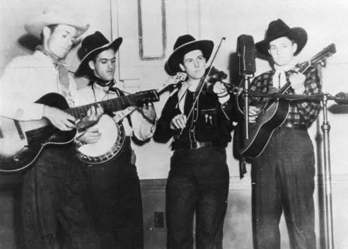 Black and white image of four men in cowboy hats standing in front of a mic playing instruments. From left to right: guitar, banjo, fiddle, guitar.