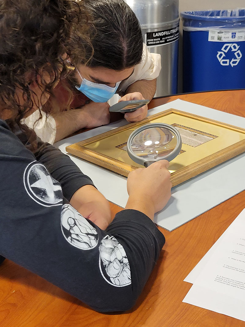Two students look at a medieval manuscript with magnifying glasses.