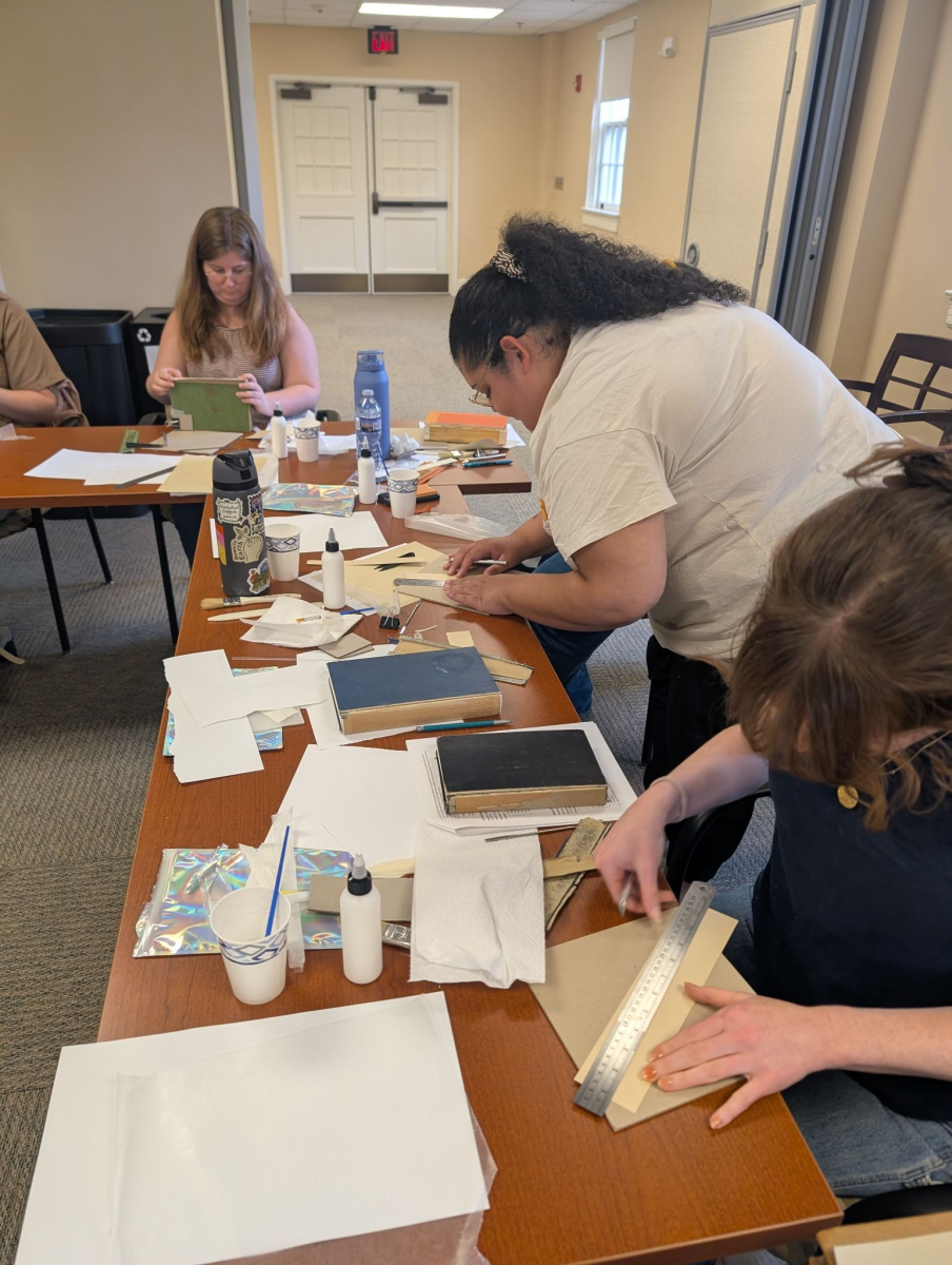 Image shows Hannah Helmey and Celine Shay working at a long table that is covered with books and book repair supplies, such as glue, papers, and brushes. They are using rulers to make measurements in preparation for spine replacement.