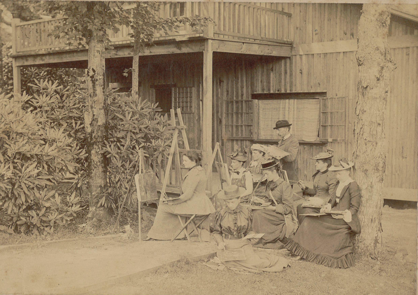 A group of people sit with easels and paint in front of a wooden building