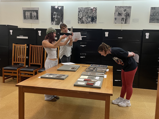 Two students and a professor examine examples of historic photography laid out on a large wooden table.