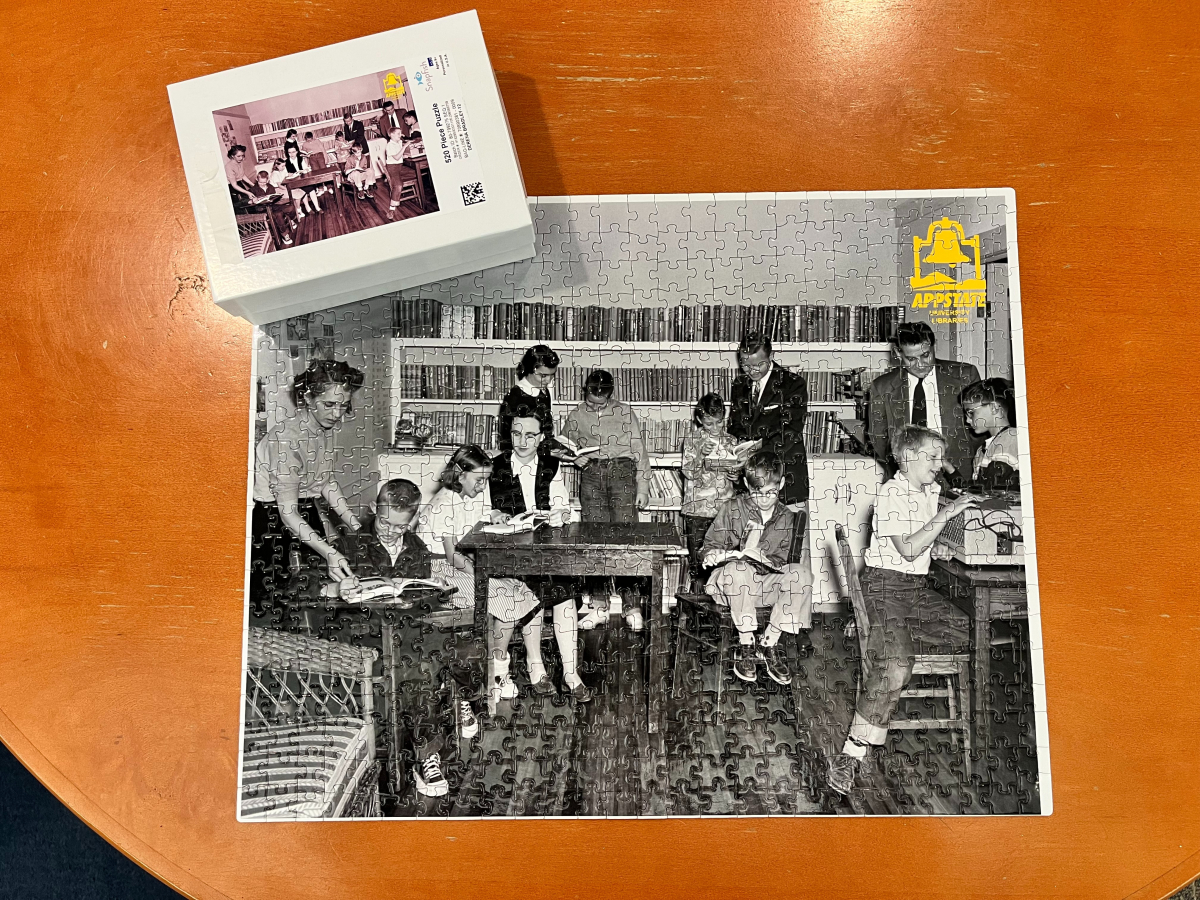 High-angle shot of a finished jigsaw puzzle titled Pic_1_Puzzle.jpg on an orange-brown wooden table. The puzzle shows a vintage, black-and-white photo of an old-fashioned classroom or library filled with children and teachers reading books. A bright yellow App State University Libraries logo sits in the top-right corner of the puzzle, and its white product box sits in the top-left.