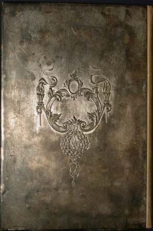 Close-up of the back cover plate of an antique book. The metallic, heavily aged silver surface features a single, centered decorative motif. The embossed design includes an elegant neoclassical crest made of scrolling acanthus leaves, flaming torches with flowing ribbons, and a detailed circular wreath dangling from the bottom center.