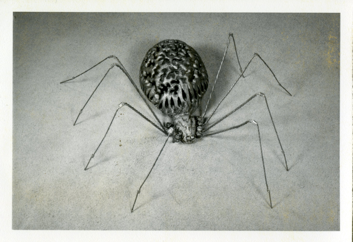 A vintage black-and-white archival photograph from a high angle showing a detailed spider sculpture. The spider has a bulbous, hollow abdomen crafted from an intricate, organic filigree pattern. Its eight long, thin legs are constructed from twisted wire, bent realistically at the joints, resting on a smooth, neutral surface.