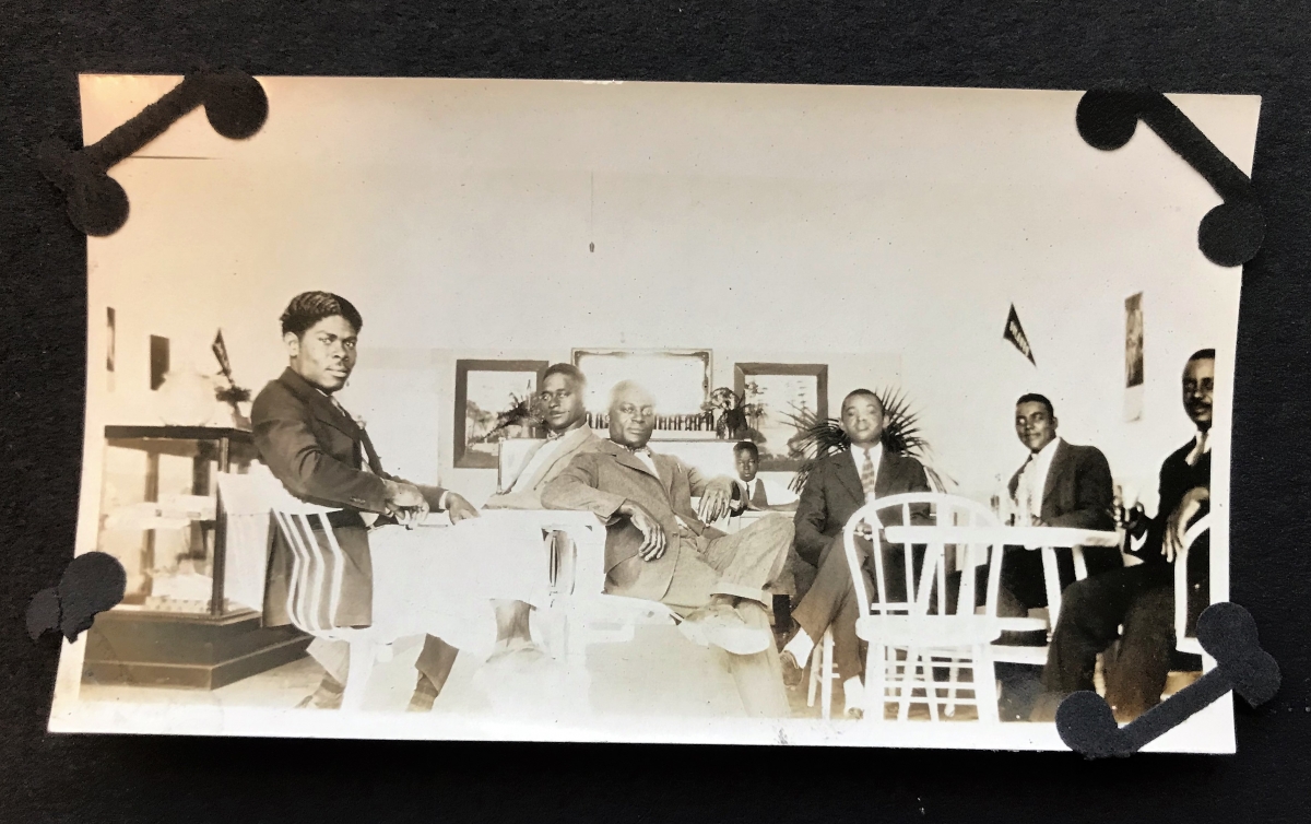 Photograph showing seven men in formal dress sitting at tables, possibly in a building of a college or university.