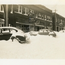 Photo depicts snowy street with vehicles and storefronts of bank, café, Farmers Hardware & Supply Co., and Quality Shoe Shop.