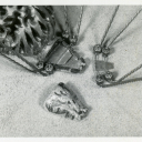 A close-up, black-and-white studio photograph showing the underside construction details of the mechanical spider. The image reveals metal plates, small hexagonal nuts, and wire attachments used to construct the legs and attach them to the body. A separate, textured, cast-metal piece—resembling a cephalothorax or head—lies detached on the surface below the main assembly.