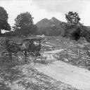 A black-and-white photograph shows a country landscape with a dirt road. A horse-and-buggy are stopped in the road, a person sitting inside the buggy.