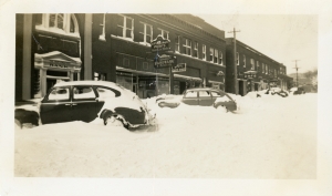 Photo depicts snowy street with vehicles and storefronts of bank, café, Farmers Hardware & Supply Co., and Quality Shoe Shop.